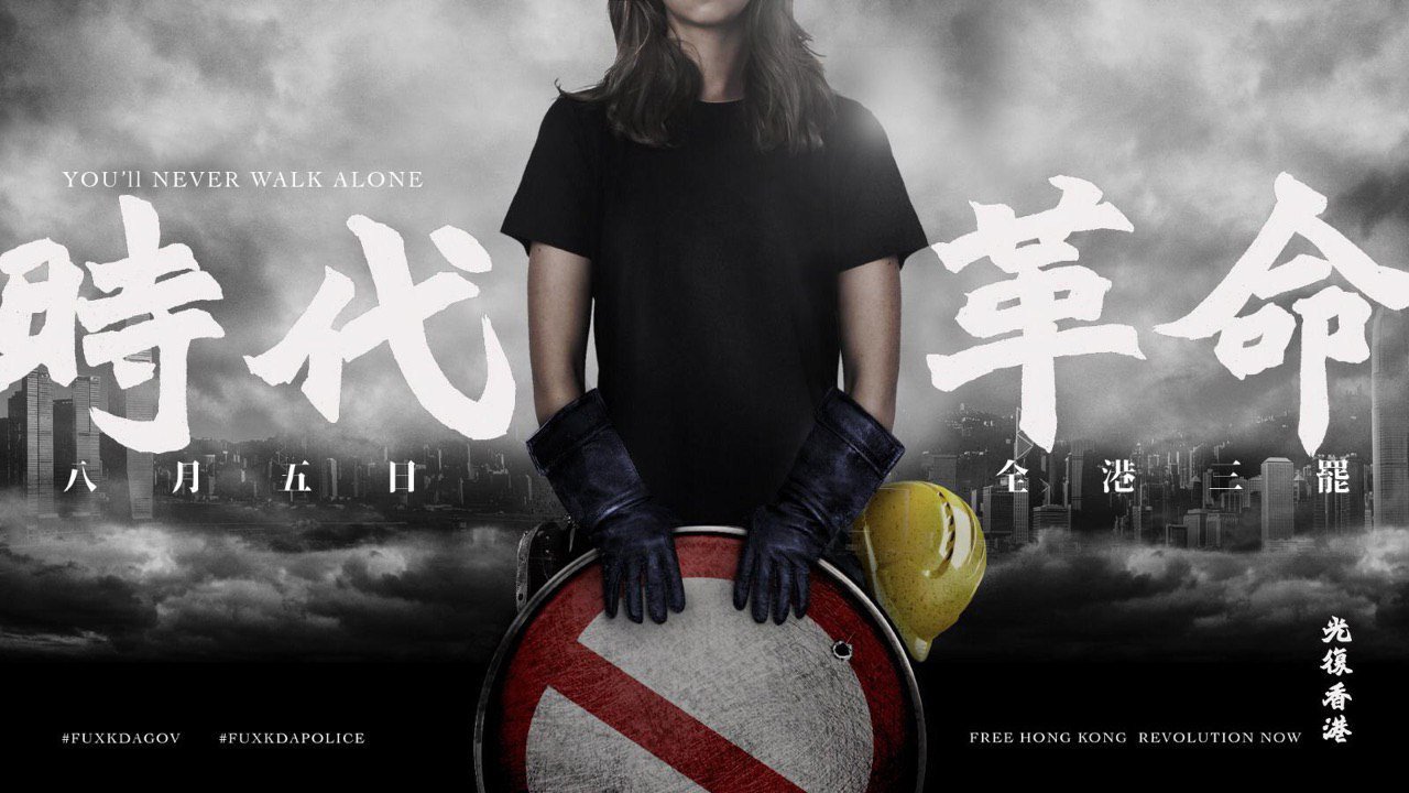 A modified photo of a young woman, her face out of shot, holding a makeshift shield made from a road sign. She is wearing a black short-sleeved tshirt and heavy-duty black gloves, with a yellow hard hat hanging from her hip. The background is a foggy black-and-white photo of the Hong Kong skyline. Text, in white, reads: You'll never walk alone (English), Revolution of Our Times (Chinese), Aug 5,  general strike across Hong Kong (Chinese). At the bottom are two hashtags, #FUXKDAGOV and #FUXKDAPOLICE, and the other half of the slogan: Free Hong Kong Revolution Now (English), Liberate Hong Kong (Chinese).
