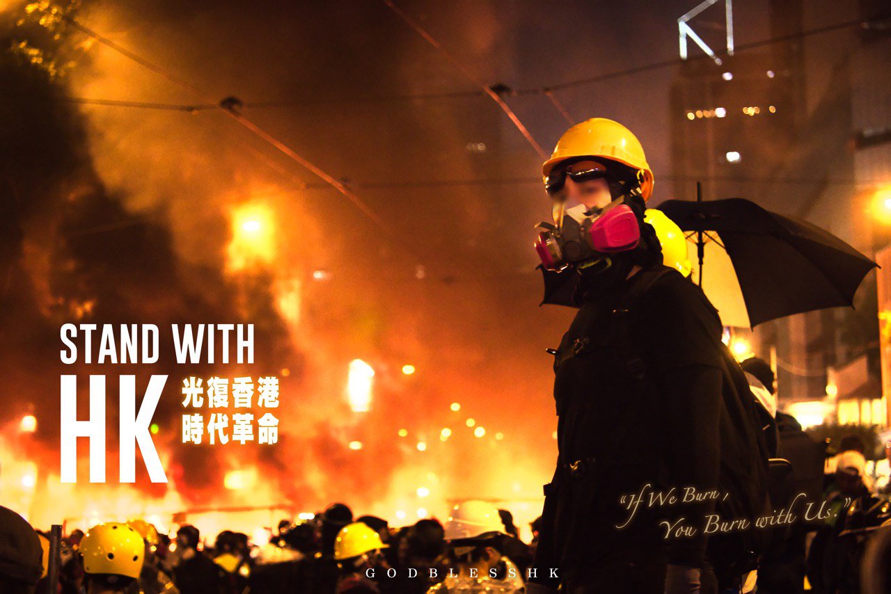 A photograph of a young frontliner in hard hat, goggles and respirator, standing against an out-of-focus backdrop of a street filled with protestors. It looks like the air is filled with gas and the glow suggests a fire in the back. Text on top, mainly English: Stand with HK. In Chinese: Liberate Hong Kong, Revolution of Our Times. English: If We Burn, You Burn With Us. God Bless HK.