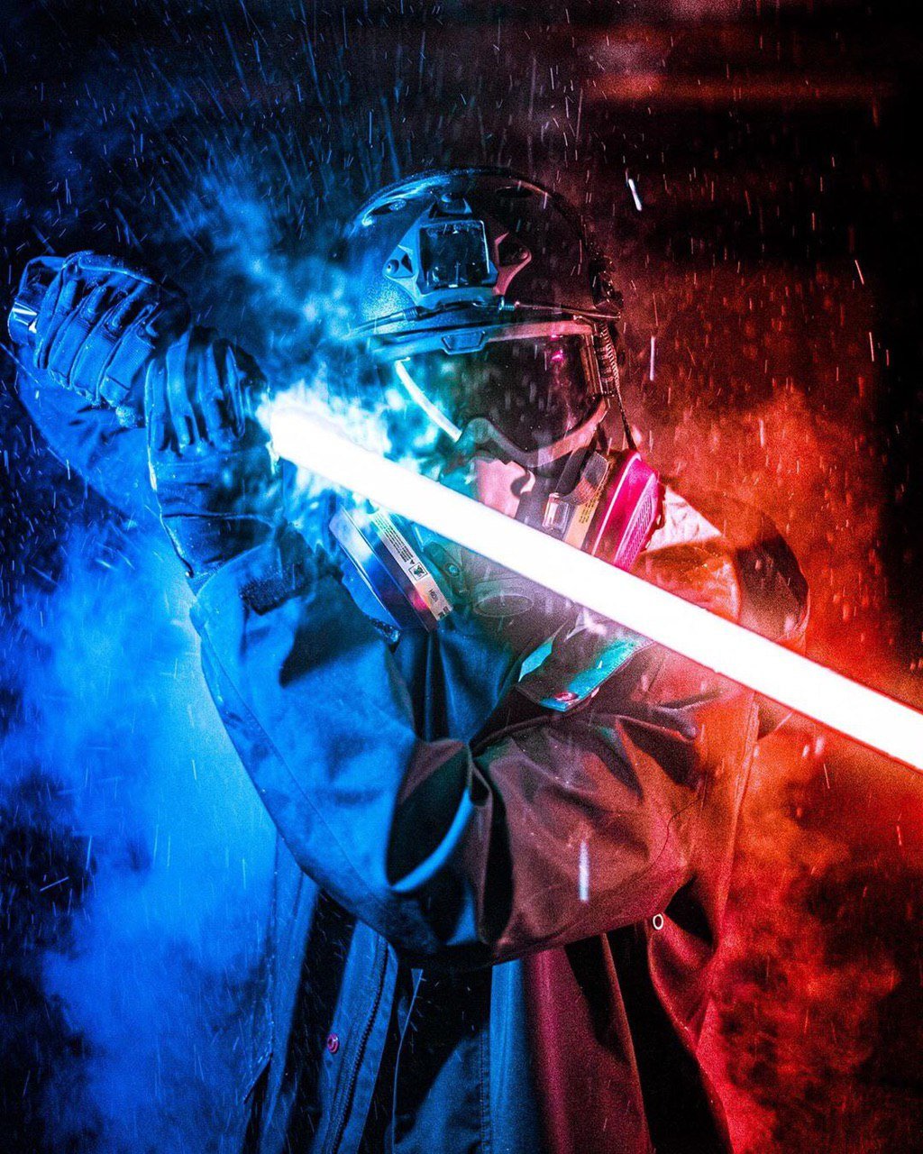 An edited photograph of a protestor in helmet, ballistic goggles and half-face respirator. They are wearing heavy-duty gloves and wielding a lightsaber. The protestor is standing in the rain against a smoky backdrop. The left side of the image is lit up in blue, while the right-hand side is in red.